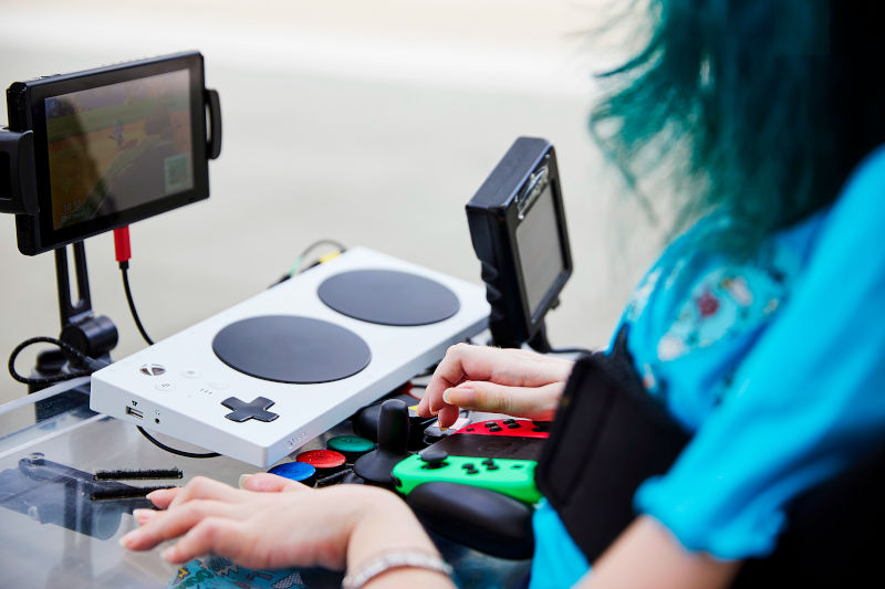 Une femme avec une amyotrophie musculaire spinale face à un petit écran de jeu vidéo et des contacteurs (gros boutons de couleurs rouge, bleu, vert) et manettes de jeu vidéo adaptées (gros boutons, boutons espacés…)