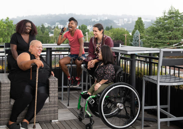 Cinq personnes handicapées de couleur, munies de cannes, de prothèses de jambes et d’un fauteuil roulant, sont assises sur un toit-terrasse, riant et partageant des histoires.