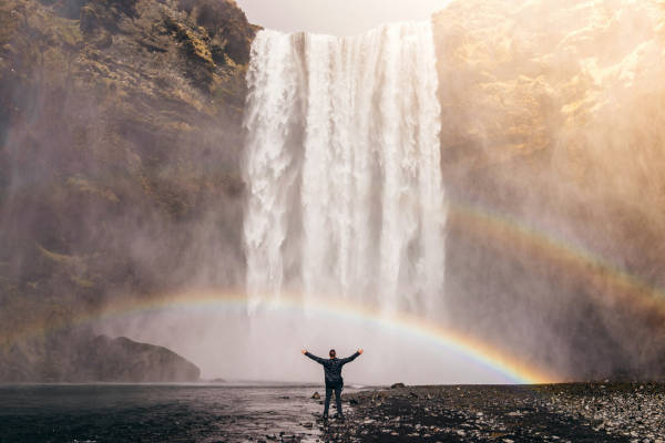 Un homme debout bras ouverts au pied et face à une cascade géante et deux arcs-en-ciel.